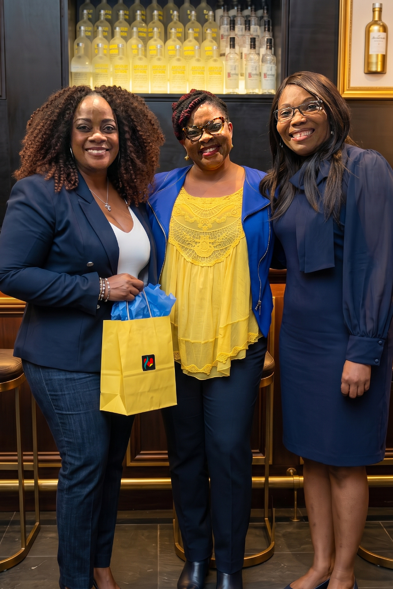 Three women smiling together indoors, two of them holding yellow gift bags.